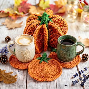 May include: A crocheted orange pumpkin with a green stem, two matching orange pumpkin-shaped coasters, a cream-colored ceramic mug with a wheat stalk design, and a speckled green ceramic mug with a leaf design on a wooden surface.