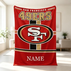 May include: A red fleece blanket featuring the San Francisco 49ers team logo in the center, with the text SAN FRANCISCO 49ERS printed above the logo in gold and white, and the word NAME printed in white at the bottom of the blanket.