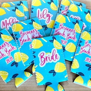 May include: A collection of blue drink koozies with a lemon pattern. The koozies have text that reads "Brienne's Beach Bach 2021", "Bride", "Lily", and "Mollie". The koozies are made of a soft, insulating material.