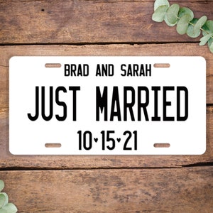 May include: White metal license plate style sign with black text that reads "Brad and Sarah Just Married 10-15-21".