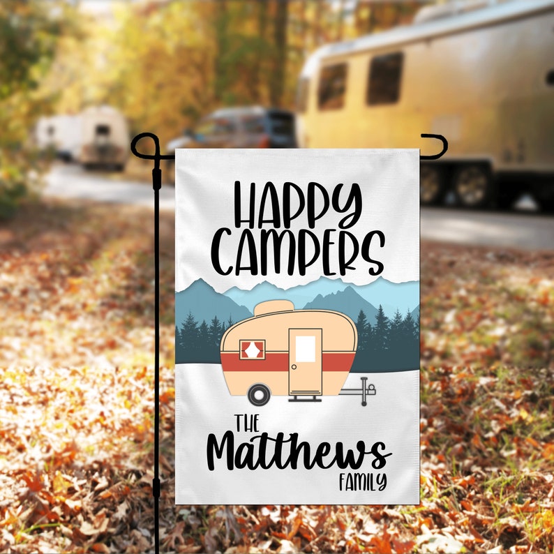 May include: A white garden flag with a cartoon camper, mountains, trees, and the text "Happy Campers The Matthews Family".