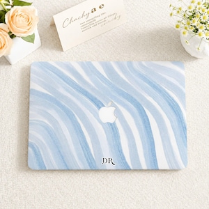 Peut inclure: Une coque rigide pour ordinateur portable avec un motif ondulé de coups de pinceau bleu clair et blanc. Le centre présente une découpe pour le logo Apple, et le bas au centre porte les initiales DR en noir avec une police serif.