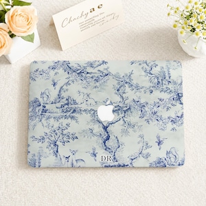 May include: A laptop cover featuring a blue and white toile de jouy pattern with illustrations of rabbits, squirrels, deer, and birds among trees and flowers. The centre displays a white Apple logo, and the letters DR are printed in black.