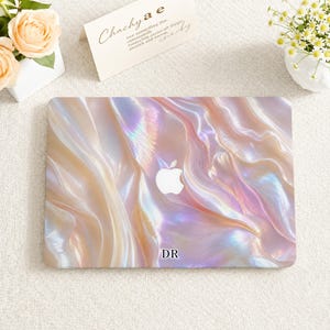 May include: A laptop skin featuring a pearlescent, iridescent finish with swirling patterns of soft pink, cream, and lavender hues. The design includes a central white Apple logo cutout and the black text DR printed near the bottom edge.