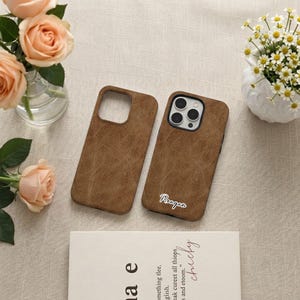 Puede incluir: Dos fundas de teléfono marrones con un acabado texturizado similar al cuero. Una de las fundas presenta el nombre Reagan en letra cursiva blanca. Las fundas tienen un recorte cuadrado para la cámara y se muestran sobre una superficie neutra.