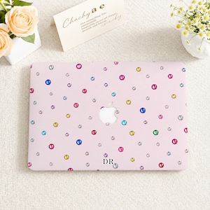 Puede incluir: Una funda rígida para portátil de color rosa claro con un patrón disperso de diamantes de imitación circulares multicolores y las letras DR impresas en negro cerca del centro inferior. La funda incluye un recorte para el logo de Apple.