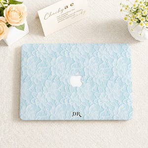 May include: A light blue laptop cover featuring a white floral lace pattern overlay. The cover displays a central cutout for the Apple logo and the black printed letters DR positioned near the bottom edge of the rectangular protective shell.