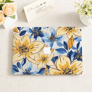 May include: A laptop hard shell case featuring a painted floral pattern in shades of yellow, navy blue, and light blue. The design includes large blossoms and leaves with a white Apple logo cutout in the center and the letters DR printed at the bottom.