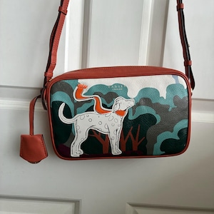 May include: A rectangular crossbody bag in burnt orange leather featuring a graphic illustration of a white spotted dog wearing an orange scarf. The background depicts stylized trees in shades of teal, dark green, and white. Includes a matching tag.