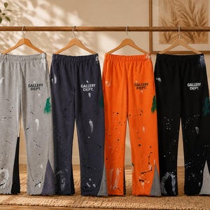 May include: Four pairs of sweatpants in gray, navy blue, orange, and black hanging on wooden hangers. Each pair features the text GALLERY DEPT. on the upper thigh and a paint splatter design with contrasting fabric panels at the bottom hems.