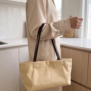 May include: A rectangular tote bag made of light beige canvas fabric with two dark brown leather shoulder straps. The bag features a single gold-toned metal snap button closure at the centre of the top opening and has a structured horizontal shape.