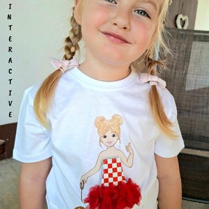 May include: A white short-sleeved t-shirt featuring a graphic of a cartoon girl with blonde hair buns, wearing a red and white checkered top with a red ruffled skirt, holding a leash attached to a small brown dog. The word INTERACTIVE is printed vertically.