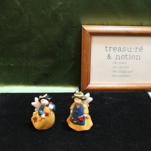 May include: Two ceramic angel figurines with white wings, blue tops, and yellow skirts. One angel holds a red item, while the other holds a red heart. A wooden frame displays the text "treasu:re & notion" with "re-used, re-cycled, re-imagined, re-claimed" below.