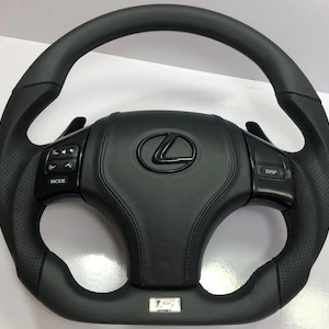 Puede incluir: Un volante de cuero negro Lexus con agarres laterales perforados, levas de cambio y botones de control integrados en los radios. El centro muestra un logotipo de Lexus negro y un emblema plateado de F Sport en la parte inferior.