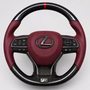 Puede incluir: Un volante Lexus con agarre de cuero rojo oscuro, detalles de fibra de carbono en la parte superior e inferior, y una franja central roja. El centro incluye un logotipo de Lexus, mientras que los radios laterales tienen botones negros.