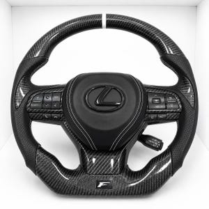 Puede incluir: Un volante Lexus negro con borde superior e inferior de fibra de carbono, agarres laterales de cuero perforado y una franja central blanca. El centro muestra un logotipo Lexus negro, costuras blancas y botones de control integrados.