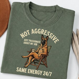 May include: An olive green t-shirt featuring a graphic of a brown German Shepherd sitting in a wooden folding chair while holding a coffee mug. The text reads: NOT AGGRESSIVE JUST PASSIONATE ABOUT MY JOB SAME ENERGY 24/7 in a cream-colored font.