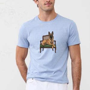 Puede incluir: Una camiseta de manga corta de color azul claro con una ilustración gráfica de un perro pastor belga malinois de color tostado y negro sentado en una silla de madera con asiento tapizado en verde. El perro está centrado en el pecho.