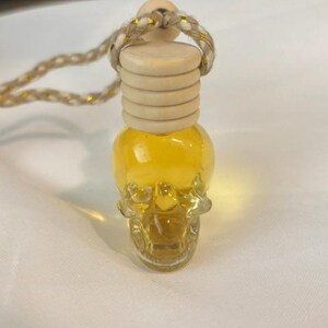 Puede incluir: Una botella de vidrio transparente con forma de calavera llena de un líquido dorado. La botella cuenta con un tapón de madera clara acanalado y está suspendida por un cordón beige trenzado con hilos dorados metálicos tejidos en el material.
