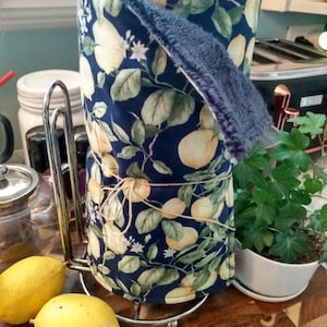 May include: A blue and green fabric kitchen towel with a lemon print is rolled up and placed on a metal paper towel holder. Two yellow lemons are in the foreground.