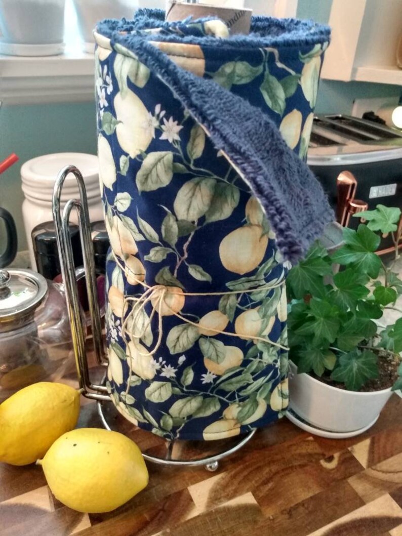 May include: A blue and green fabric kitchen towel with a lemon print is rolled up and placed on a metal paper towel holder. Two yellow lemons are in the foreground.