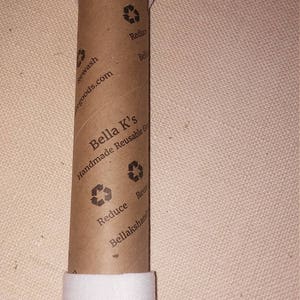May include: A brown cardboard tube with white tape wrapped around it. The tube has the text "Bella K's Handmade Reusable Goods" printed on it, along with a recycling symbol and the words "Reduce, Reuse, Bellakshandmadegoods.com".