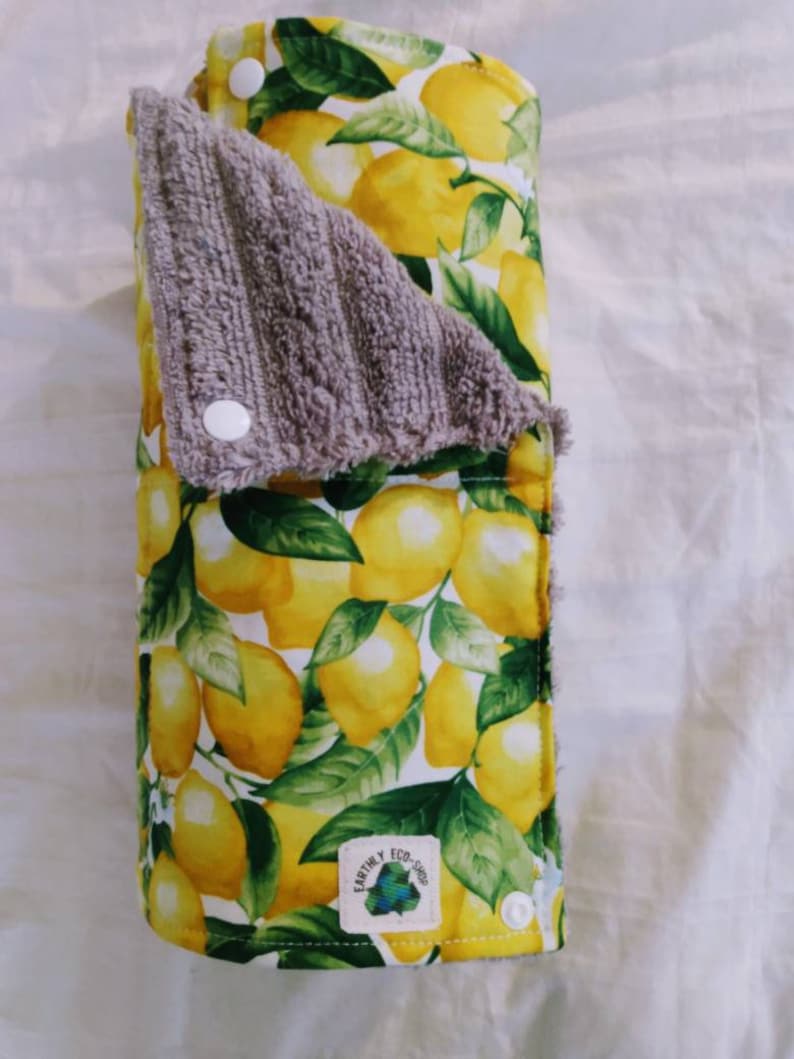 May include: A reusable paper towel made of fabric with a lemon print. The towel is rolled up and has a gray terry cloth side. The towel has a white label with the text "Earthly Eco Shop".