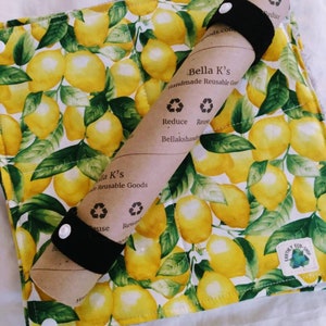 May include: A brown paper roll with black elastic bands wrapped around it. The paper roll has text that reads "Bella K's Handmade Reusable Goods" and "Reduce Reuse Bellakshands". The roll is on top of a white fabric with a lemon print. The fabric has a green and yellow lemon pattern and the text "Earthy Eco-Shop".