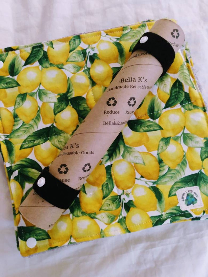 May include: A brown paper roll with black elastic bands wrapped around it. The paper roll has text that reads "Bella K's Handmade Reusable Goods" and "Reduce Reuse Bellakshands". The roll is on top of a white fabric with a lemon print. The fabric has a green and yellow lemon pattern and the text "Earthy Eco-Shop".