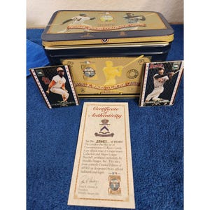 Puede incluir: Una caja de almacenamiento rectangular de metal azul oscuro con detalles dorados y gráficos de jugadores de béisbol, acompañada de dos tarjetas de colección y un certificado de autenticidad con el texto Set No. 25657 of 49,900.
