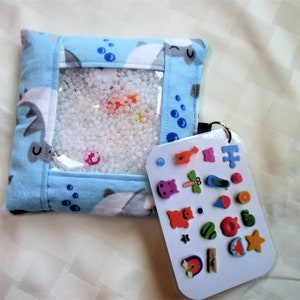 May include: A blue fabric sensory bag with a clear plastic window filled with white beads. A white card with a variety of colorful shapes and objects is attached to the bag.