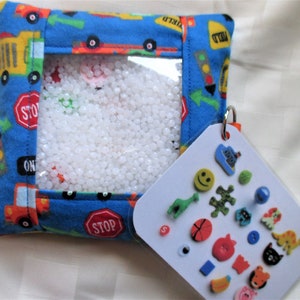 May include: A blue fabric sensory bag with a clear window filled with white beads. The bag has a red zipper and a red and yellow truck pattern. A white card with colorful buttons attached to the bag with a silver ring.