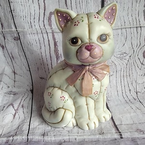 May include: A ceramic cat figurine in a seated position, featuring a cream-colored body with painted pink floral patterns and a mauve-colored muzzle. The cat has large brown eyes, long eyelashes, and a mauve ribbon tied in a bow around its neck.