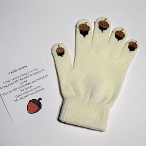 May include: A white knitted fingerless glove with five brown acorn appliques. The glove is accompanied by a card with the text "5 Little Acorns" and a short poem about a squirrel and acorns.
