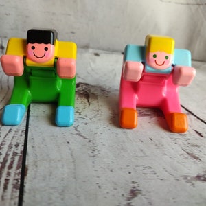 May include: Two colorful plastic figurines of people sitting with their legs crossed. The figurine on the left is wearing a yellow shirt, green pants, and blue shoes. The figurine on the right is wearing a blue shirt, pink pants, and orange shoes.