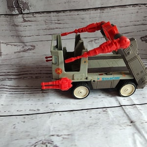May include: A grey and red toy truck with the word "STARCOM" printed on the side. The truck has two red cannons on the back and two red cannons on the front.