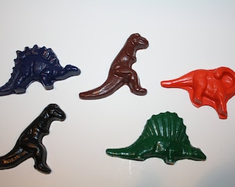 May include: Five colorful dinosaur-shaped crayons in various colors: dark blue, brown, red, black, and green. The crayons are arranged on a white surface, showcasing their distinct shapes and hues.