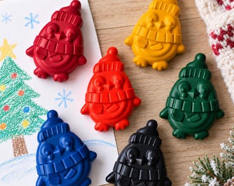 Peut inclure: Un ensemble de cinq crayons de cire en forme de pingouin, déclinés en rouge, jaune, vert, bleu et noir. Chaque crayon présente un motif moulé de pingouin portant un bonnet et une écharpe. Un crayon repose sur un papier blanc avec un sapin.