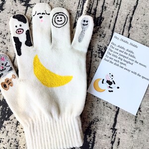 May include: White knitted finger puppet glove with a yellow crescent moon, a cow, a cat, a dog, a smiley face, and a spoon. The glove is on a textured surface with a card that reads "Hey, Diddle, Diddle. Hey, diddle, diddle, The cat and the fiddle, The cow jumped over the moon; The little dog laughed To see such sport, And the dish ran away with the spoon."