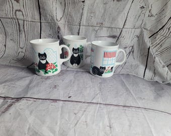 Peut inclure: Trois tasses en céramique blanche ornées d'illustrations de chats noirs. Une tasse montre un chat près de fleurs rouges, une autre affiche un chat assis, et la troisième représente un chat près d'une fenêtre avec un auvent rayé rouge et blanc.