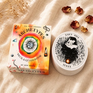 May include: The Wild Unknown Archetypes deck and guidebook box features a colourful watercolour design with a central eye illustration. Beside it sits a circular card from the deck displaying a black and white illustration titled The Cave IIXXX.