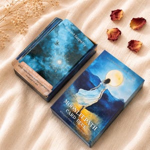 May include: A deck of cards titled Moonlit Path Card Deck with an illustration of a person in a blue dress under a full moon. Beside it, an open box displays a card titled Radiant Geometry featuring a hummingbird and geometric star patterns.