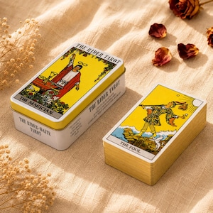 Op de afbeelding: Een Rider-Waite tarotdeck in een wit metalen blik met de kaart De Magiër op het deksel, vergezeld door een aparte stapel kaarten met de kaart De Dwaas bovenop. De kaarten hebben goudkleurige randen en liggen op een beige textielstof.
