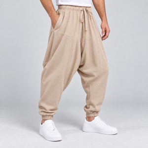 May include: Beige drop-crotch trousers featuring an elasticated waistband with a drawstring closure, side pockets, and gathered elastic cuffs at the ankles. The trousers have a relaxed, loose-fitting silhouette with vertical seam detailing.