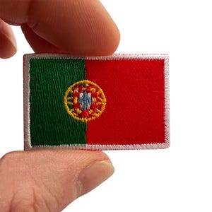 May include: A rectangular embroidered patch featuring the flag of Portugal with a green vertical band on the left and a larger red vertical band on the right, centred with a yellow, red, and blue emblem, finished with a white stitched border.