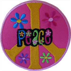 Può includere: Una toppa ricamata circolare con sfondo rosa e un simbolo di pace dorato. La parola Peace è scritta in lettere nere stilizzate al centro, decorata con piccoli fiori colorati nelle tonalità del blu, del rosa e del verde sulla superficie.