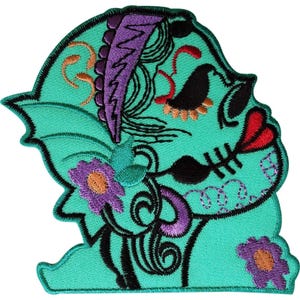 May include: An embroidered patch featuring a stylised profile of a creature in vibrant teal. The design includes a purple jagged crest, black eye and mouth details, bright red lips, and purple flowers with orange centres, all outlined in black thread.
