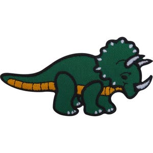 May include: An embroidered iron-on patch featuring a green triceratops dinosaur with a yellow underbelly and white horns. The dinosaur has a large frill with white accents and is outlined in a thick black border against a plain white background.