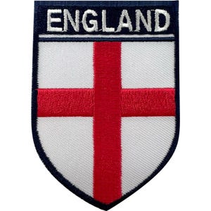 May include: A shield-shaped embroidered patch featuring a white background with a red cross. The top section is a dark blue horizontal band with the word ENGLAND stitched in white capital letters. The entire patch has a dark blue border.