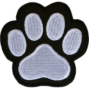 May include: An embroidered patch in the shape of an animal paw print. The design features a solid black background with four oval-shaped toe pads and one large, triangular-shaped central pad, all stitched in a light grey thread with a visible texture.